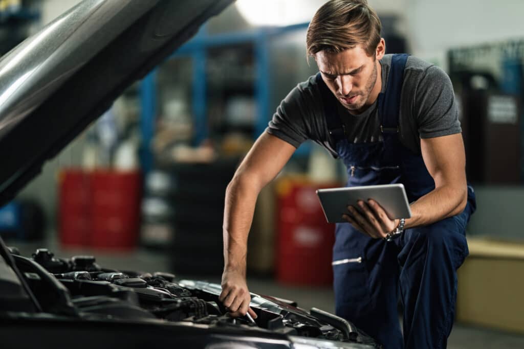 Young auto repairman working on car engine while using digital tablet in repair shop.